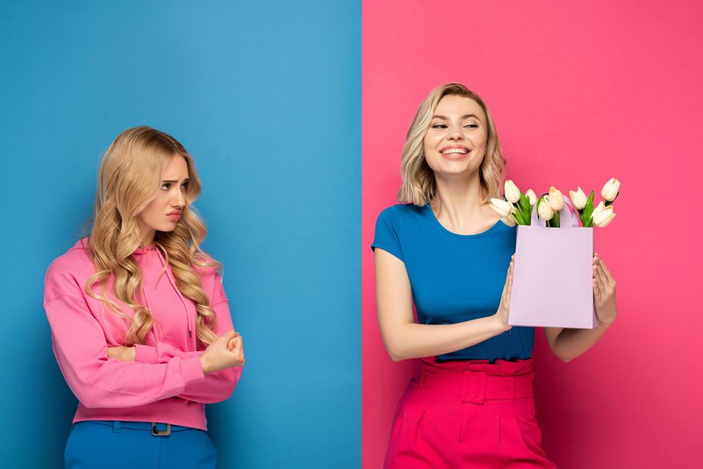 boos blond meisje op zoek naar gelukkig zus met boeket op roze en blauwe achtergrond - Foto, afbeelding
