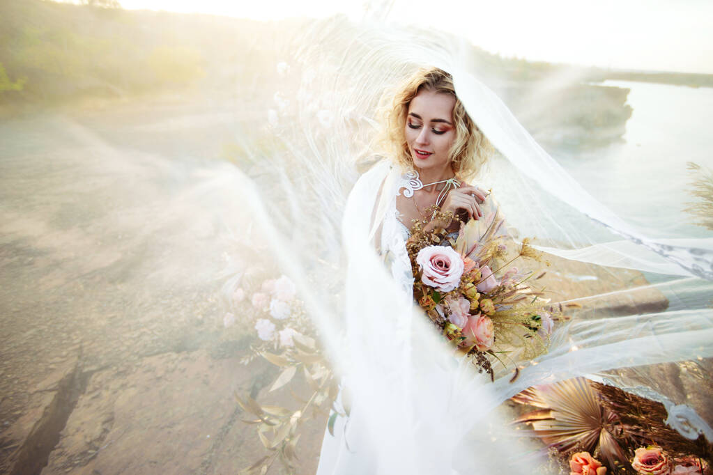 La novia cerca de la decoración de la boda en una ceremonia en un acantilado de roca cerca del agua al atardecer. Velo volando del viento
 - Foto, imagen