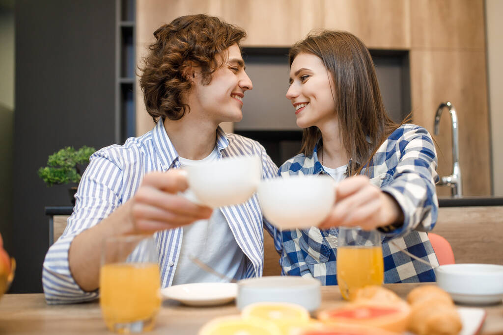 家でのコーヒータイム。かわいい若いカップルは自宅のキッチンでコーヒーを飲む、カップを保持. - 写真・画像