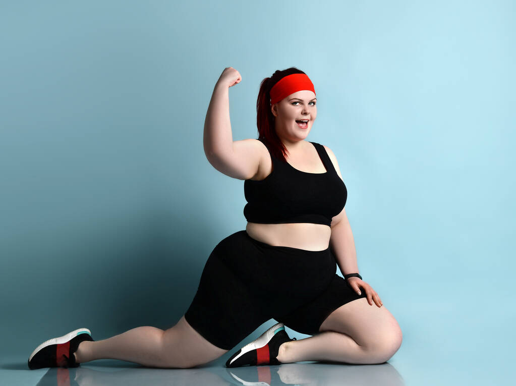 Femme rousse taille plus en bandeau rouge, haut noir, shorts, baskets. Montrant les muscles, debout sur les genoux sur fond bleu
 - Photo, image