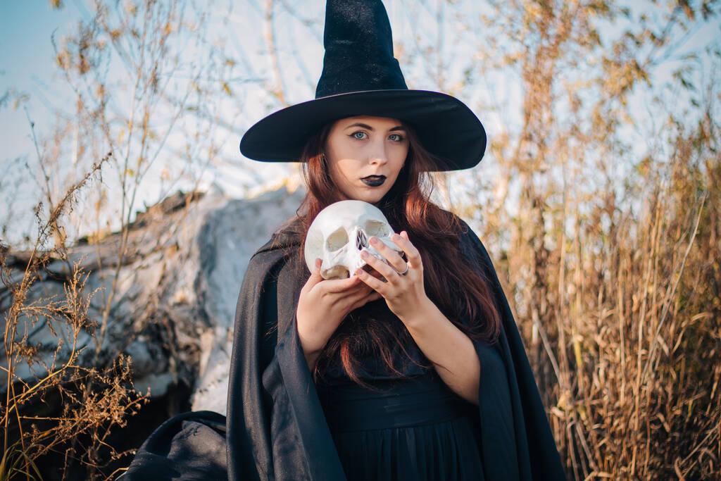 Egy fiatal boszorkány sápadt bőrrel és fekete ajkakkal fekete kalapban, ruhában és esőkabátban. Egy halott ember koponyáját fogja. Ősz, kidőlt fa és magas, száraz fű. Halloween, varázslat, fantázia. - Fotó, kép