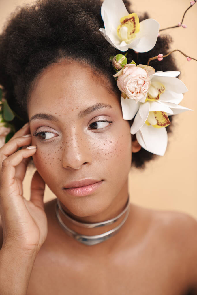 Porträt einer jungen halbnackten sommersprossigen afrikanisch-amerikanischen Frau mit Blumen im Haar, isoliert auf beigem Hintergrund - Foto, Bild