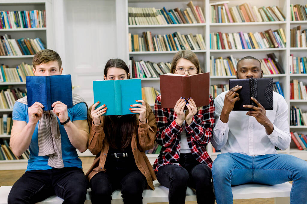 Four multiethnical friends students, two boys and two girls in casual wear, hiding faces behind open books and looking at camera. Group of students with books in modern university library - Photo, Image