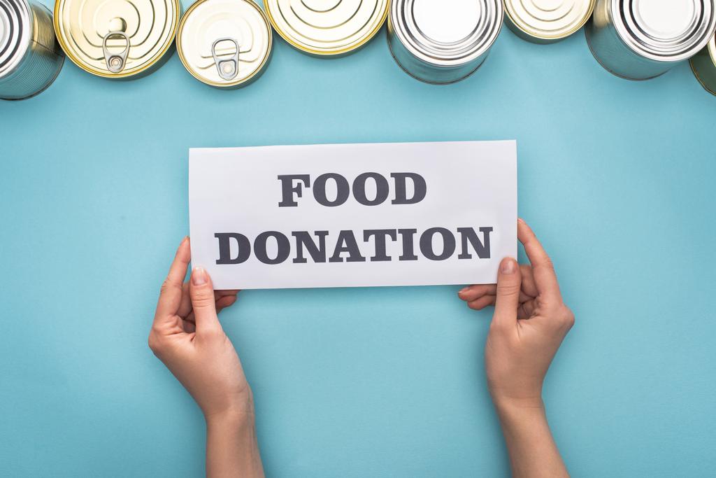 bijgesneden weergave van vrouw met kaart met voedsel donatie belettering in de buurt van blikken op blauwe achtergrond - Foto, afbeelding