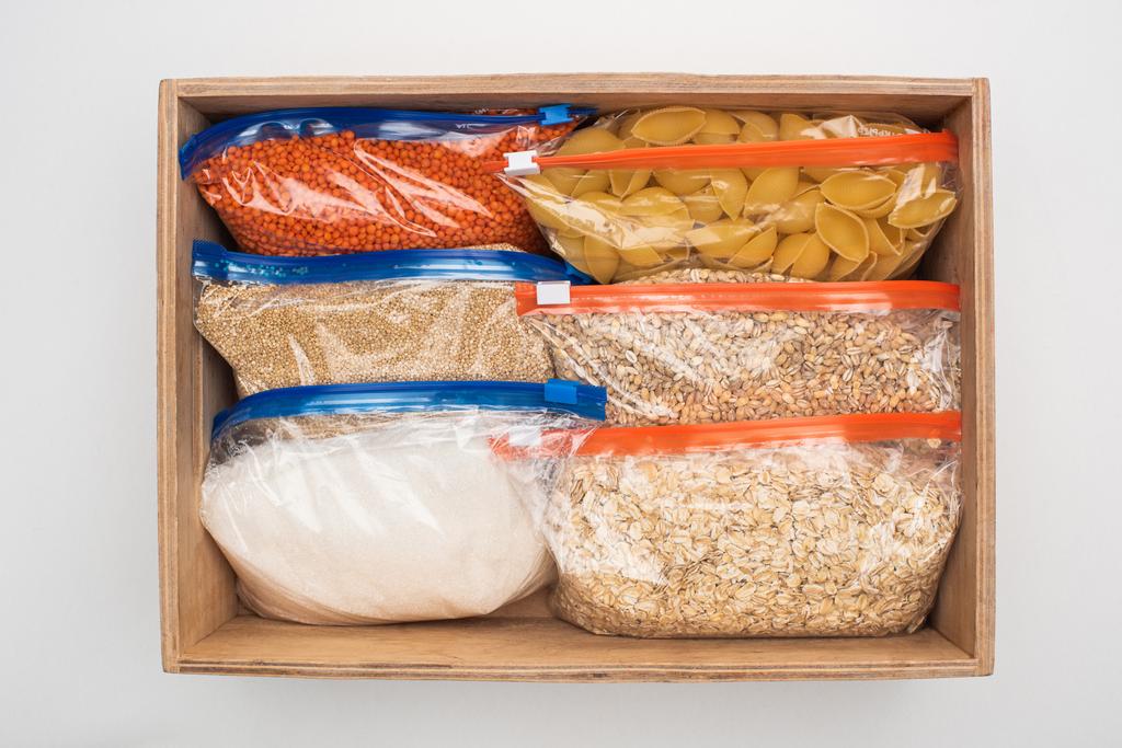 vrchní pohled na těstoviny a krupice v sáčcích na zip v dřevěné krabici na bílém pozadí, koncept dárcovství potravin - Fotografie, Obrázek