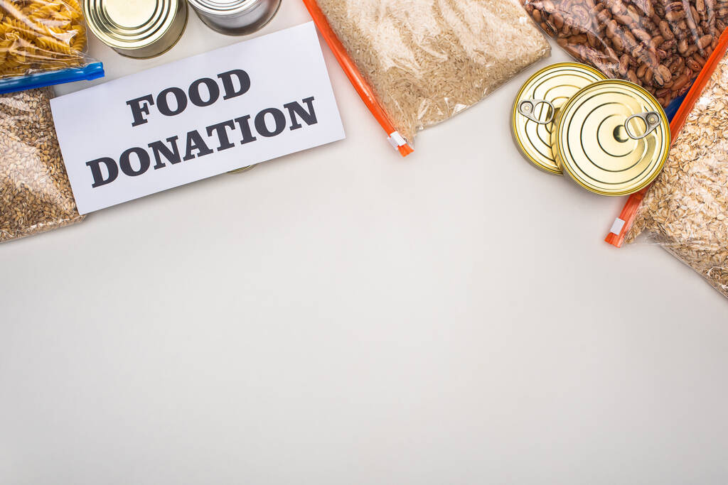 вид зверху банок і крупи в мішках на блискавці біля картки з харчовим донорством на білому тлі
 - Фото, зображення