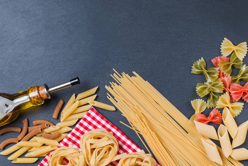 Different types of pasta and bottle of olive oil on a black slate background - Photo, Image