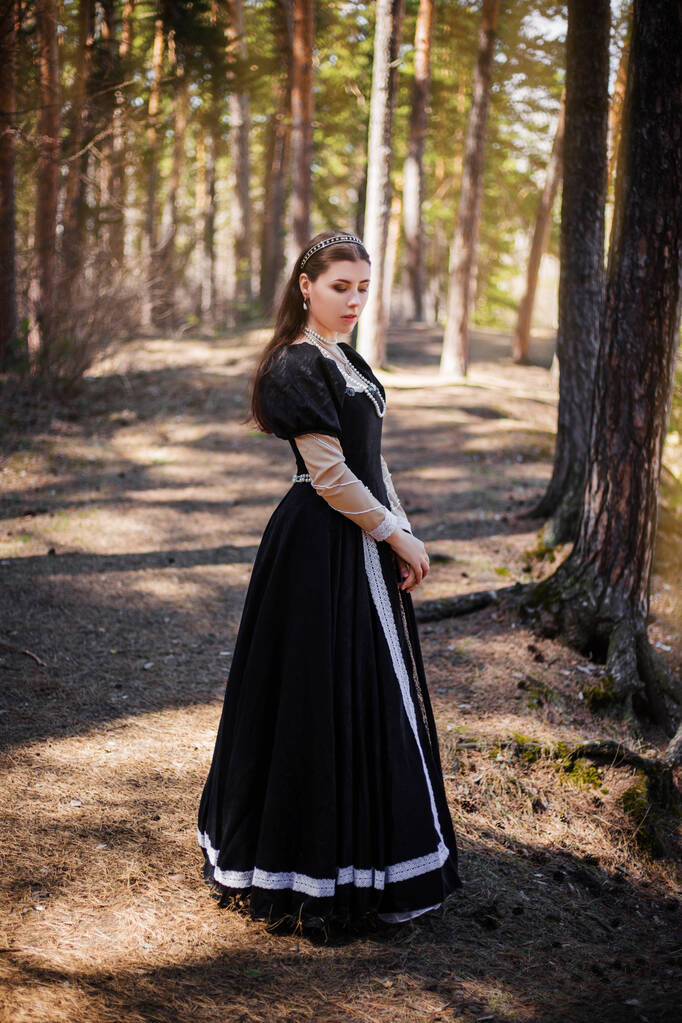 Fiatal, gyönyörű nő fekete középkori ruhában sétál a borús fenyőerdőben.. - Fotó, kép