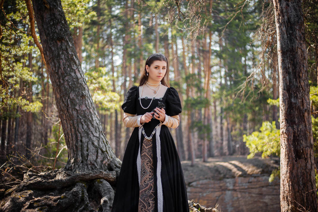 Mladá, krásná žena v černých středověkých šatech s ocelovou růží v ruce, kráčející lesem. Atraktivní dívka v gotických šatech. - Fotografie, Obrázek
