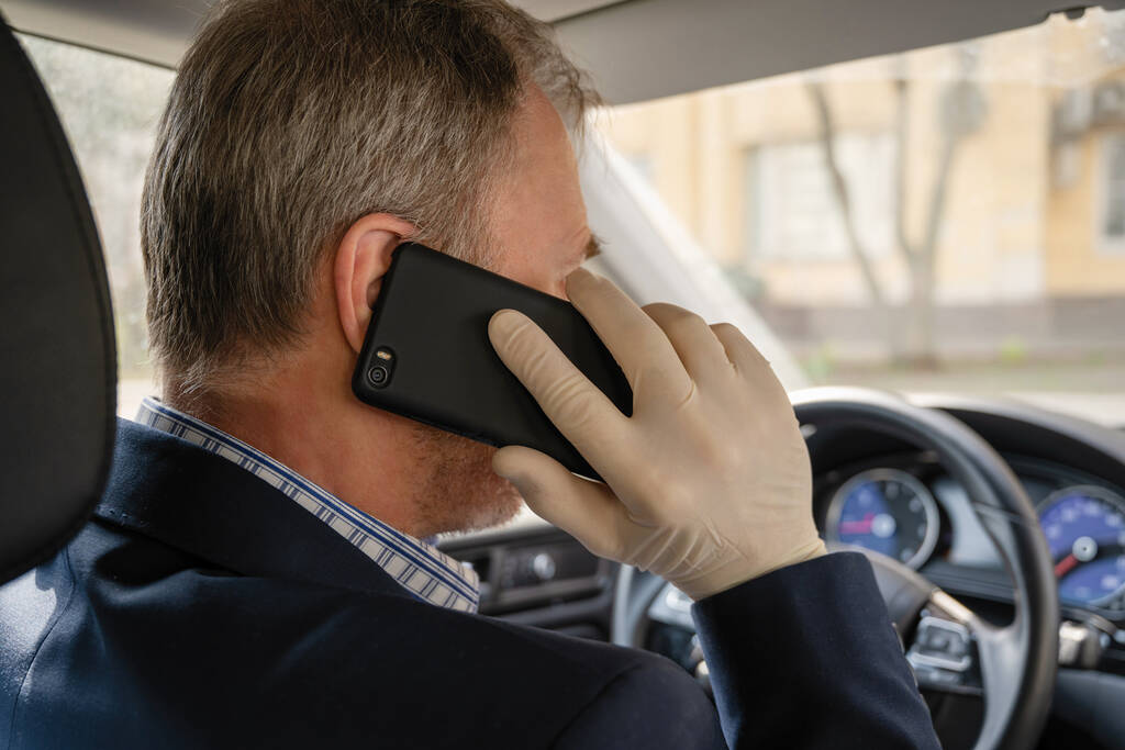 homme d'âge moyen appelant au téléphone dans des gants en caoutchouc tout en conduisant une voiture. protection contre les bactéries et les virus pendant la conduite d'une voiture
. - Photo, image