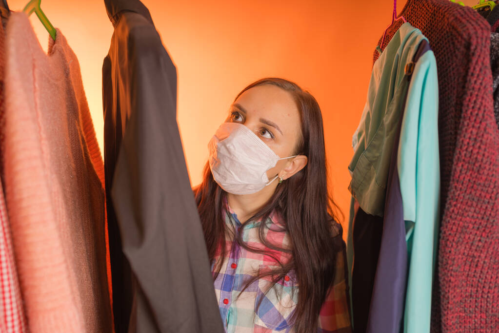 Close up de jovem mulher em máscara médica à procura do que escolher roupas no guarda-roupa
. - Foto, Imagem