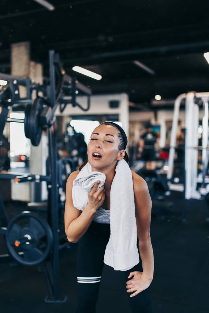 Young exhausted woman relaxing in fitness gym after successful workout wipes sweat from the forehead with white towel. - Photo, Image