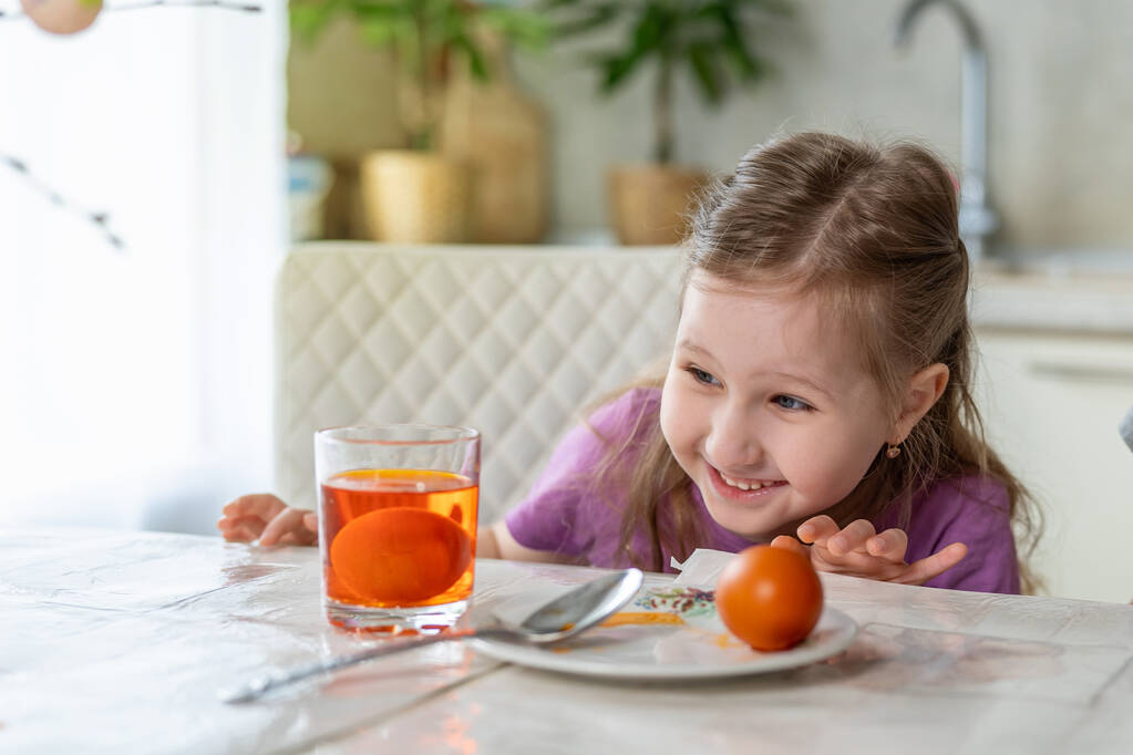 イースターの準備だ。かわいい女の子がテーブルに座って卵をペイントします。子供は卵が着色されるまで待ち、驚きと喜びで着色を見る. - 写真・画像