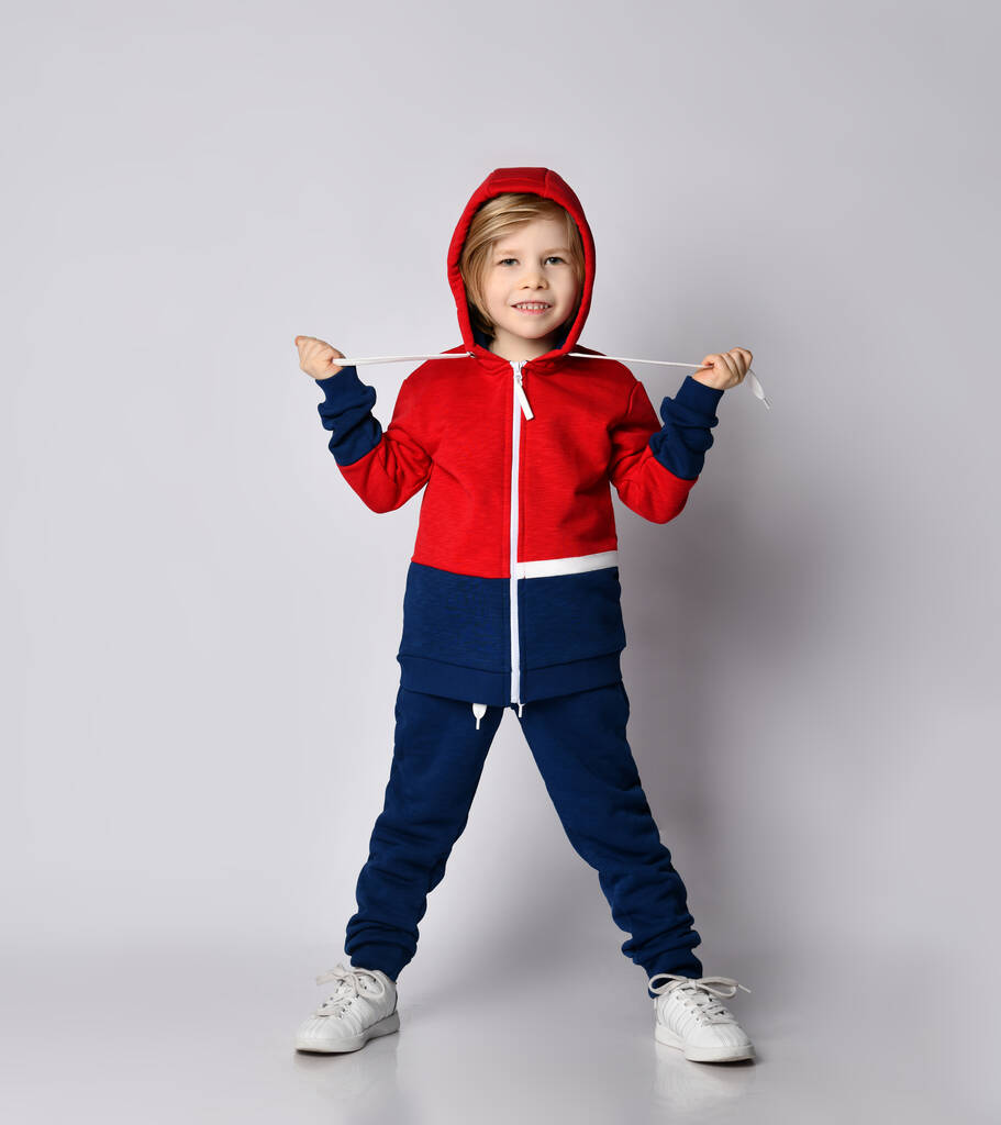 Παιχνιδιάρικο ξανθό αγόρι με μπλε και κόκκινο σπορ κοστούμι ποζάρει με κουκούλα στο κεφάλι. - Φωτογραφία, εικόνα