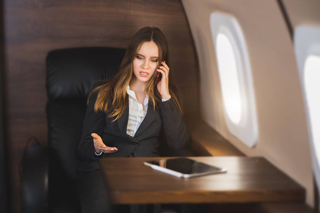 Attraente giovane donna d'affari caucasica nervosa sta parlando al telefono con un'espressione facciale irritata, gesticolare, seduta su un aereo privato. Business, concetto di emozioni. - Foto, immagini