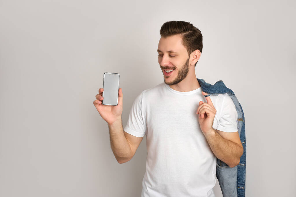 Alegre hipster en camiseta blanca aislado sobre fondo gris, presentando teléfono inteligente, mirándolo y sosteniendo una chaqueta de mezclilla sobre su hombro, sonriendo, sintiéndose positivo
. - Foto, imagen