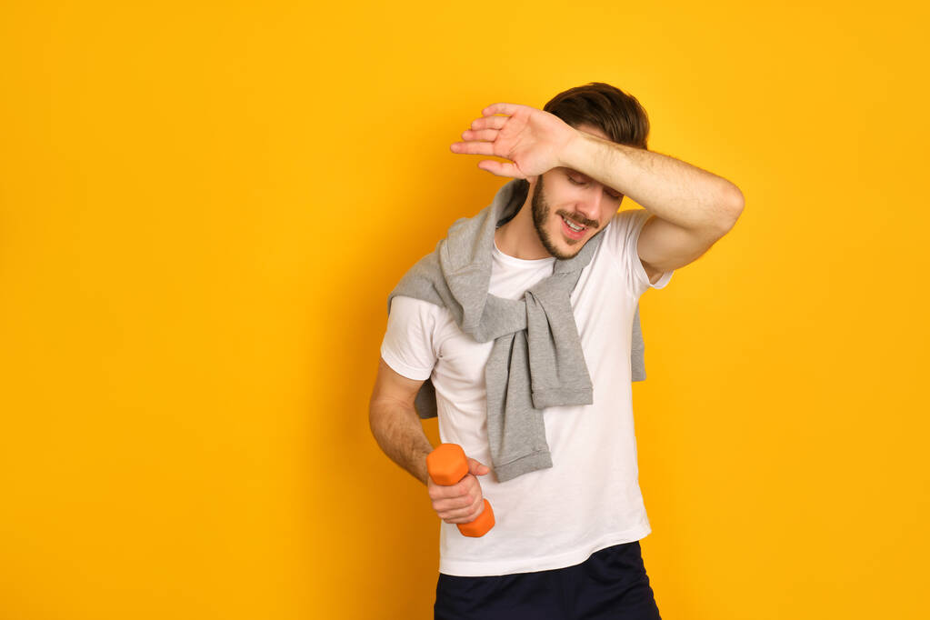 Un joven deportista con barba en camiseta blanca y suéter beige sobre fondo amarillo, riéndose, sosteniendo una mancuerna en una mano, cuidando su salud, sintiéndose cansado, se limpia la frente
 - Foto, imagen