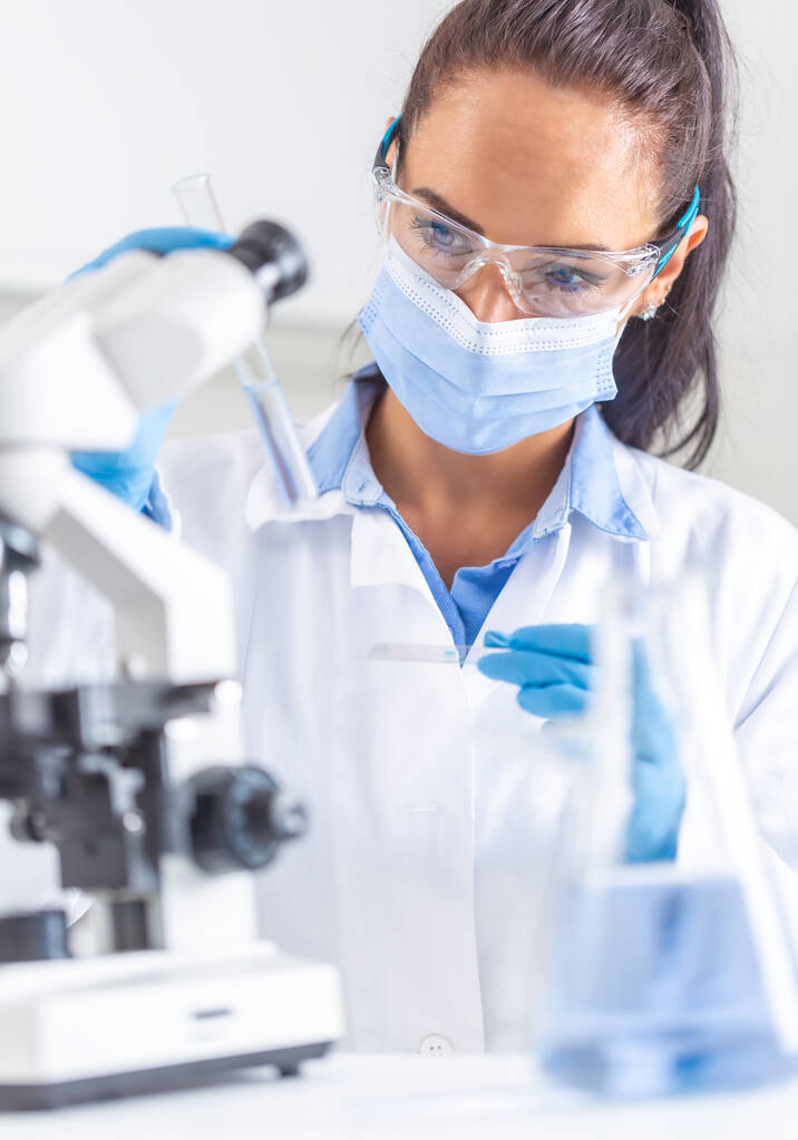 Mulher de casaco branco, óculos de proteção e máscara facial olha para o tubo de teste em uma mão, segurando uma lâmina de microscópio na outra mão. Conceito de Coronavirus COVID-19
. - Foto, Imagem