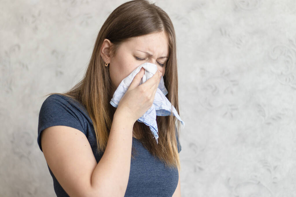Sick woman with handkerchief sitting at home with flue, virus, cold, seasonal allergy symptom. Young woman blows her nose or sneezing in a blue cloth handkerchief. - Photo, Image