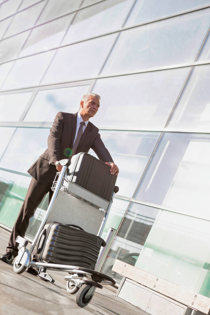 Портрет зрелого бизнесмена, толкающего багажную тележку с чемоданом в аэропорту
 - Фото, изображение