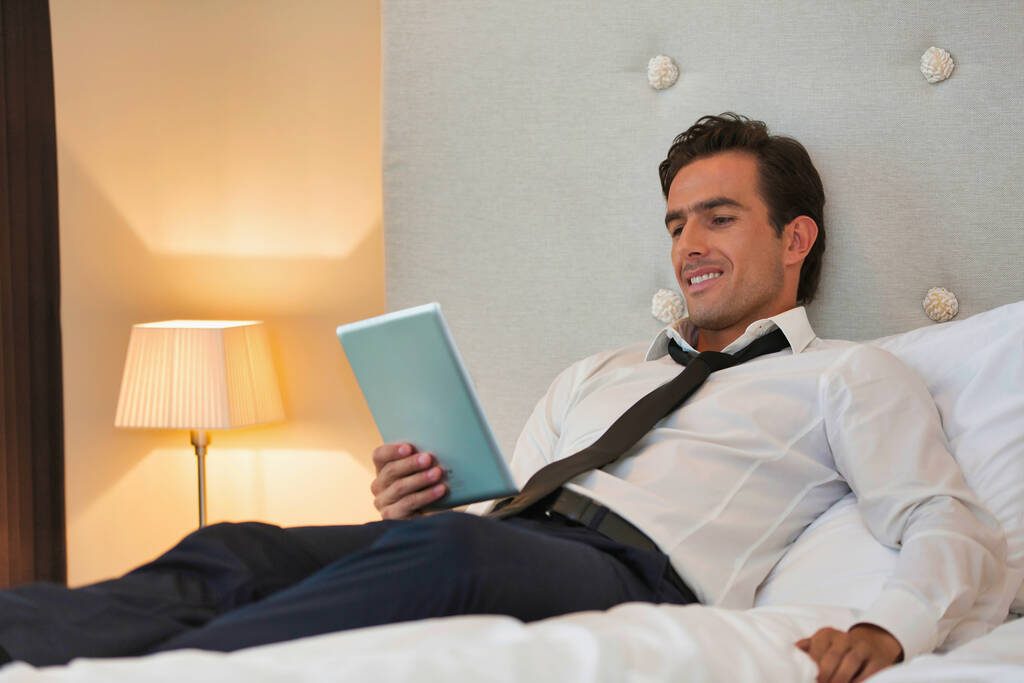 Kypsä liikemies makaa sängyllä, kun käytät digitaalista tablettia kotona
 - Valokuva, kuva