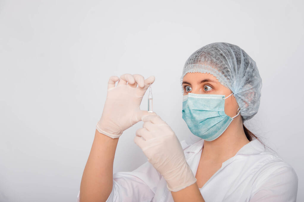 Крупный план ампулы вакцины в руках женщины-врача. Лаборант в белом халате, медицинской шляпе, маске и перчатках, держит в руках лекарство. Концепция поиска коронавирусной вакцины
. - Фото, изображение