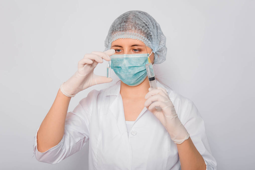 Ο γιατρός κρατάει μια σύριγγα και έναν αμπούλα με ένα εμβόλιο. Νεαρή γυναίκα με λευκό παλτό, με ιατρική μάσκα, καπέλο και γάντια σε λευκό φόντο. Εργαστηριακός ιατρός. Έννοια αναζήτησης εμβολίων του Coronavirus. - Φωτογραφία, εικόνα