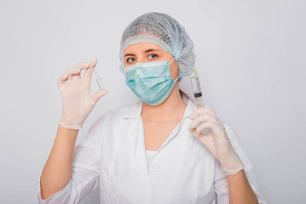 Ο γιατρός κρατάει μια σύριγγα και έναν αμπούλα με ένα εμβόλιο. Νεαρή γυναίκα με λευκό παλτό, με ιατρική μάσκα, καπέλο και γάντια σε λευκό φόντο. Εργαστηριακός ιατρός. Έννοια αναζήτησης εμβολίων του Coronavirus. - Φωτογραφία, εικόνα