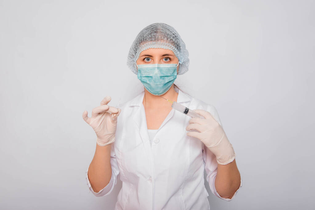 Lékař drží injekční stříkačku a ampulku s vakcínou. Mladá žena v bílém plášti, v lékařské masce, klobouku a rukavicích na bílém pozadí. Laboratorní lékař. Koncept hledání koronavirové vakcíny. - Fotografie, Obrázek