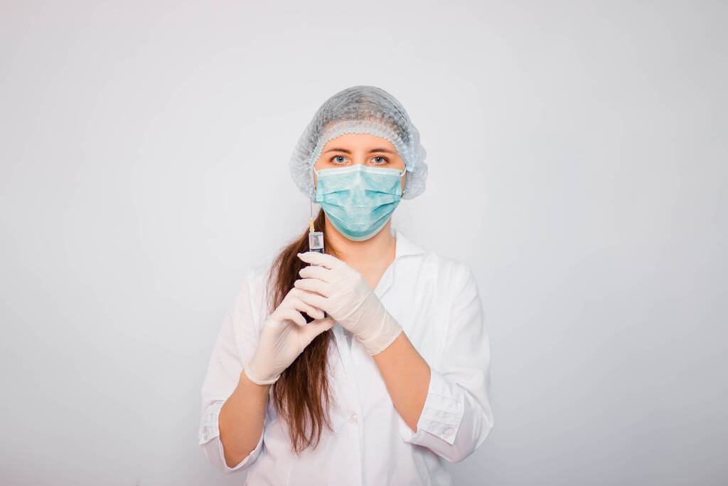 Γυναίκα γιατρός σε λευκό ιατρικό παλτό, μάσκα, καπέλο και γάντια κρατά μια σύριγγα με το φάρμακο στα χέρια της. Στέκεται πάνω σε λευκό φόντο. Έννοια αναζήτησης εμβολίων του Coronavirus. - Φωτογραφία, εικόνα