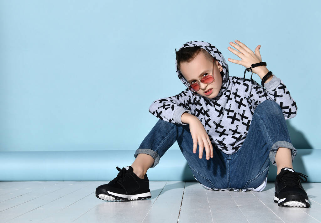 Teenager σε γυαλιά ηλίου, μπλέ τζιν, φούτερ, μαύρο βραχιόλι και sneakers. Σηκώθηκε το χέρι, κάθεται στο λευκό πάτωμα. Μπλε φόντο - Φωτογραφία, εικόνα
