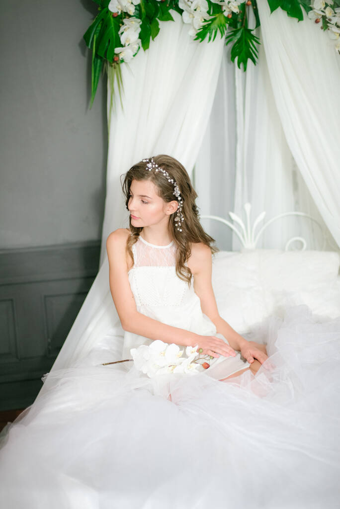 Atrakcyjna młoda dziewczyna w romantycznym białym dressonie białe łóżko z baldachimem i białymi orchideami w rękach. Piękno i moda - Zdjęcie, obraz
