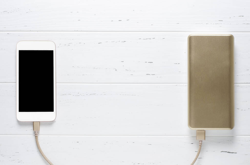 白い木製の背景にポータブルパワーバンクとスマートフォン。フラットトップビュー。スペースのコピー - 写真・画像
