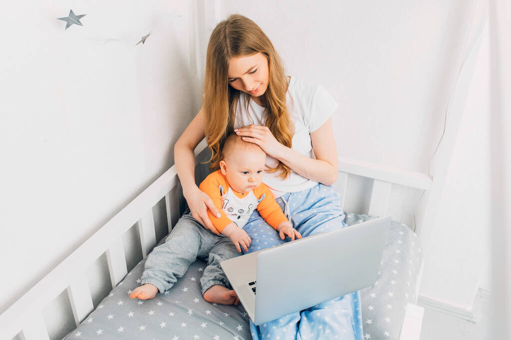 小さな子供を持つパジャマを着た若い母親は、ポータブルラップトップでベッドの上に座っています。隔離における遠隔作業の概念 - 写真・画像
