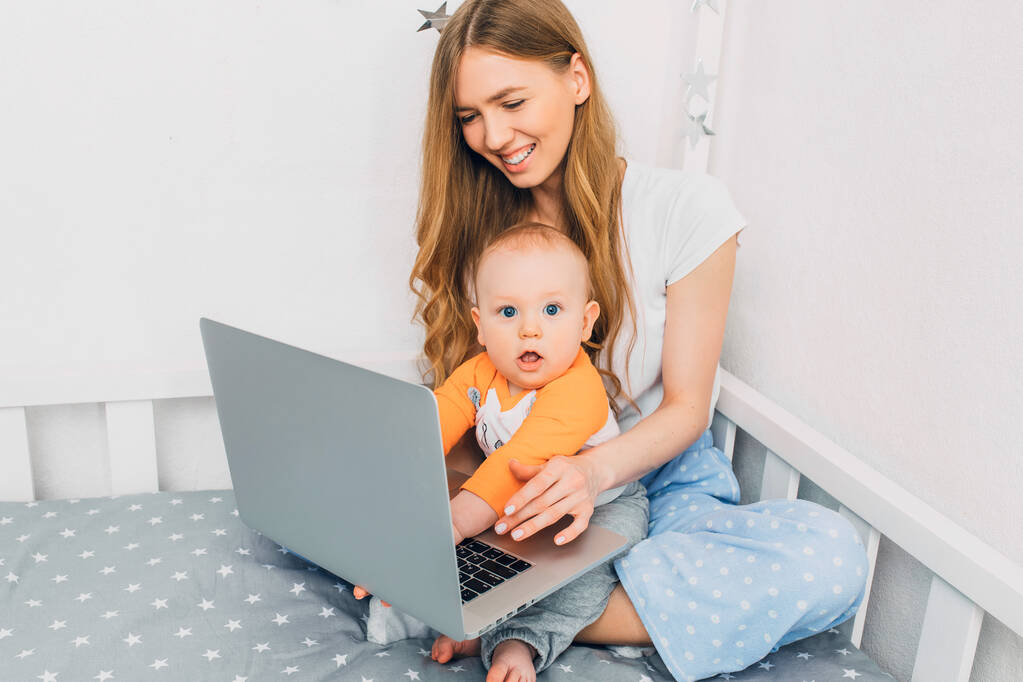 Una joven madre en pijama con un niño pequeño, sentada en la cama, con un portátil. El concepto de trabajo remoto en cuarentena
 - Foto, imagen