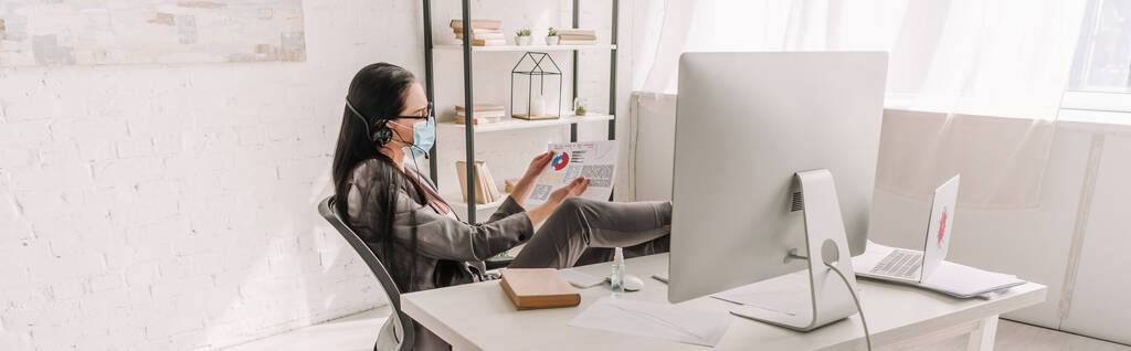 панорамний знімок бізнес-леді в медичній масці, дивлячись на папери біля столу з комп'ютерним монітором під час роботи вдома
 - Фото, зображення