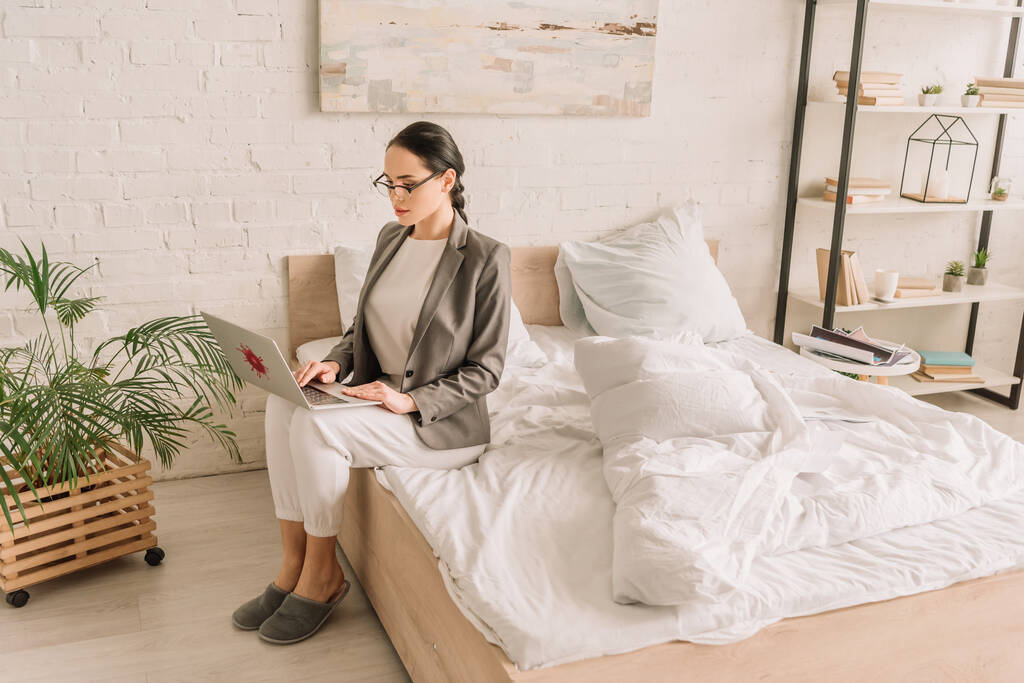 young businesswoman in blazer over pajamas using laptop while sitting on bed - Photo, Image