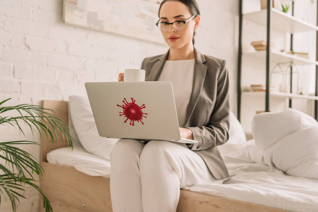 приваблива бізнес-леді в блістері над піжамою, використовуючи ноутбук і тримаючи чашку кави, сидячи на ліжку
 - Фото, зображення