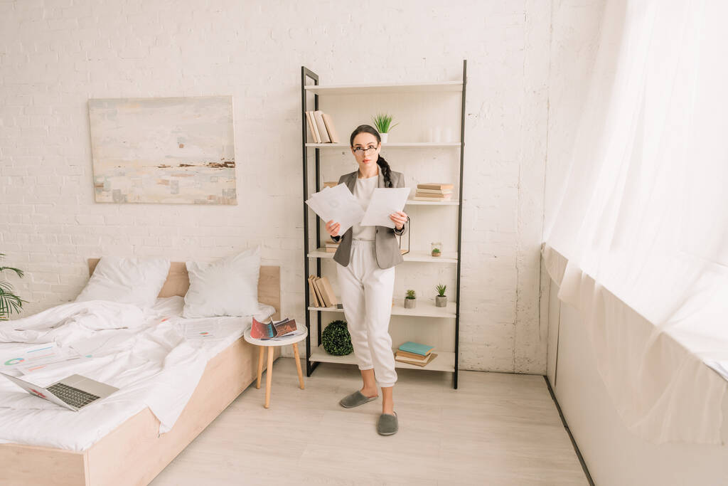 młoda bizneswoman w marynarce nad piżamą trzymając papiery patrząc na aparat fotograficzny w sypialni - Zdjęcie, obraz