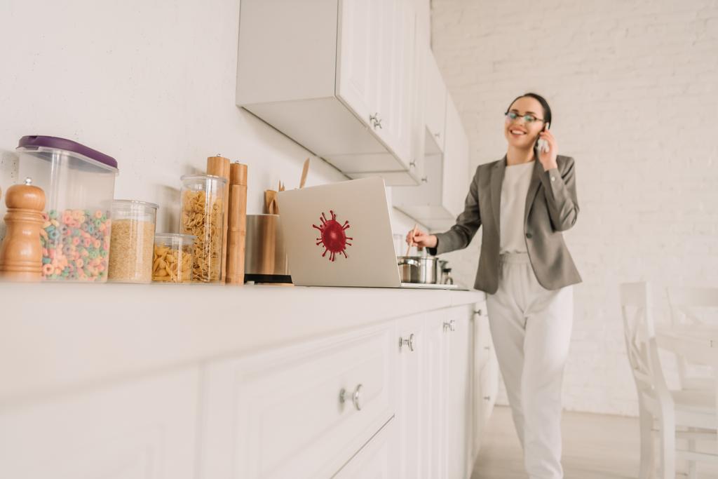 selektiver Fokus der Geschäftsfrau im Blazer über dem Schlafanzug Kochen und Reden auf dem Smartphone in der Küche in der Nähe von Laptop mit Coronavirus-Bakterien-Aufkleber - Foto, Bild