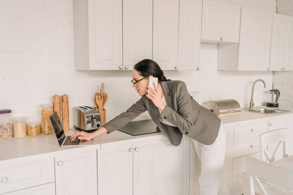 Geschäftsfrau im Blazer über Pyjama spricht auf Smartphone und benutzt Laptop in der Küche - Foto, Bild