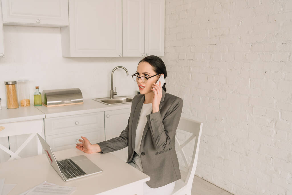 young businesswoman in blazer over pajamas talking on smartphone while working in kitchen near laptop - Photo, Image