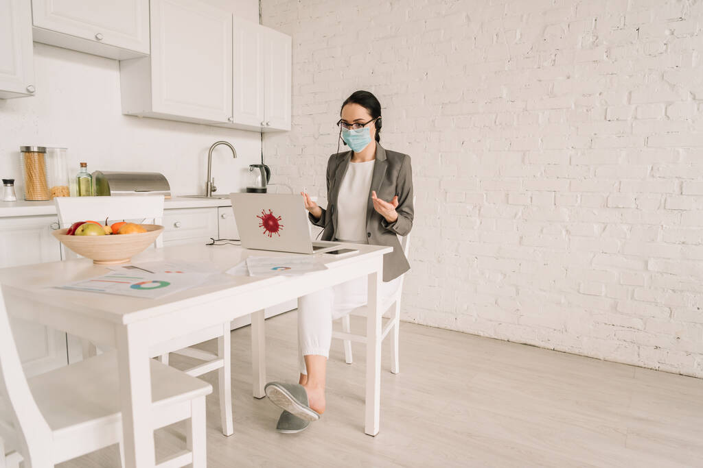 деловая женщина в медицинской маске и пиджаке на жесты во время работы на кухне рядом с ноутбуком
 - Фото, изображение