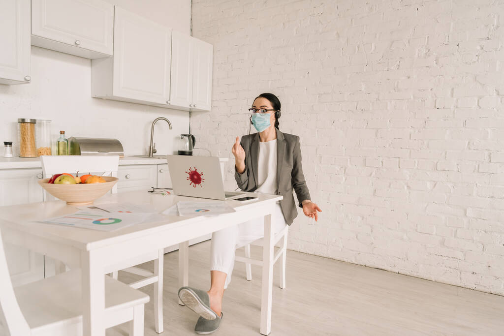 femme d'affaires en colère dans le masque médical et blazer sur pyjama montrant majeur tout en travaillant dans la cuisine près de l'ordinateur portable
 - Photo, image
