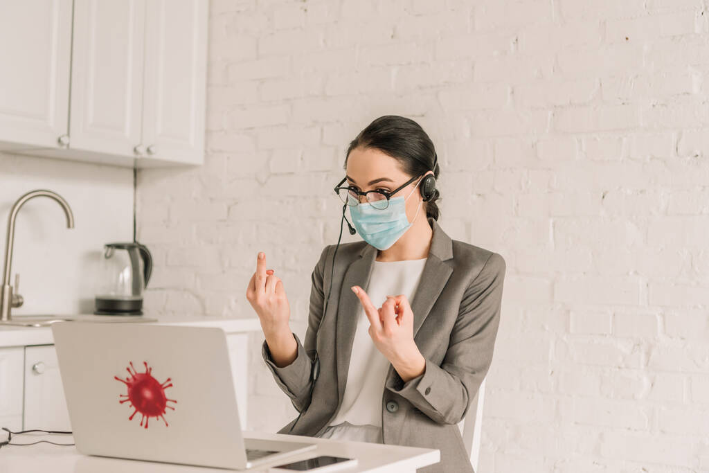 сердитая деловая женщина в медицинской маске и наушниках, показывающая средний палец во время работы на кухне возле ноутбука
 - Фото, изображение