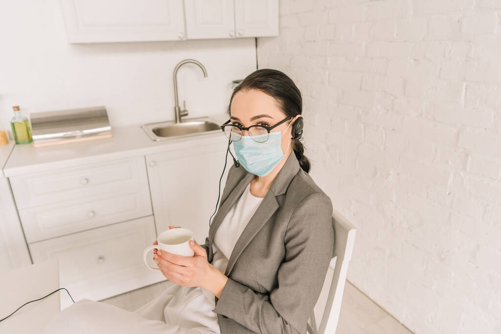 mladá podnikatelka v lékařské masce, sluchátka a blejzr drží šálek kávy v kuchyni - Fotografie, Obrázek