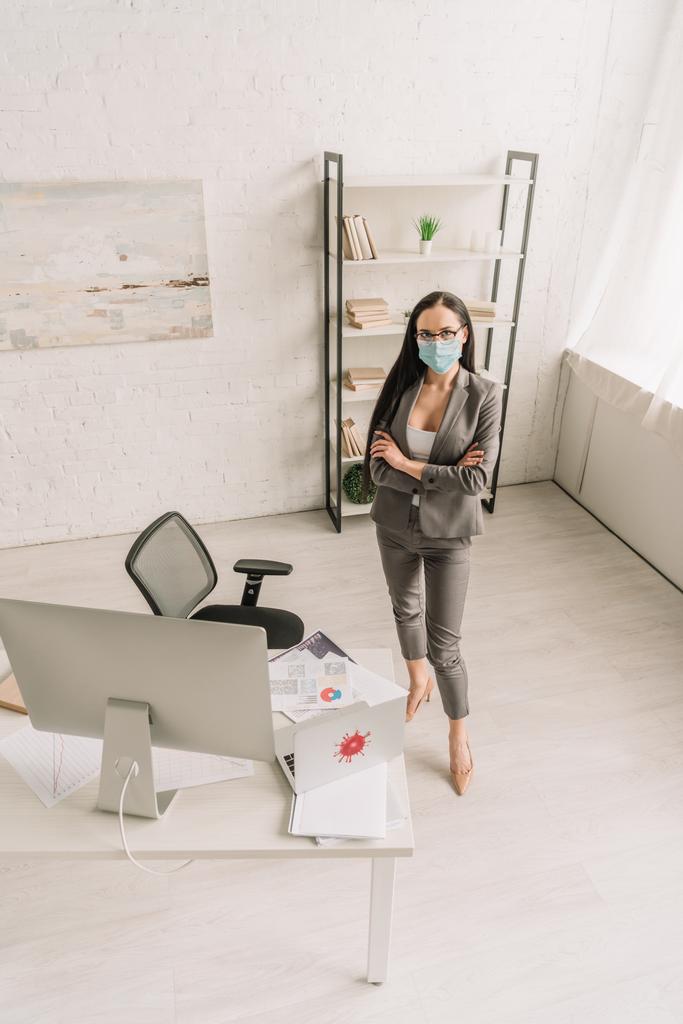 Hochwinkelaufnahme einer Geschäftsfrau in formeller Kleidung und medizinischer Maske, die mit verschränkten Armen am heimischen Tisch mit Computern steht - Foto, Bild