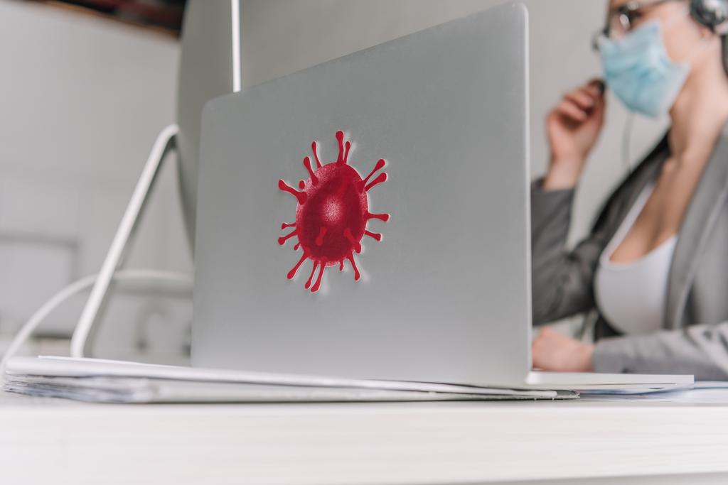 селективный фокус ноутбука с наклейкой коронавируса бактерий рядом с бизнесвумен в медицинской маске работает на дому
 - Фото, изображение