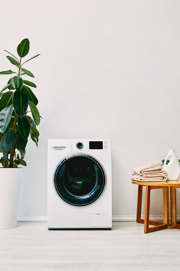 zöld növény közelében mosógép és fa dohányzóasztal törölközővel és mosószer üveg a fürdőszobában  - Fotó, kép