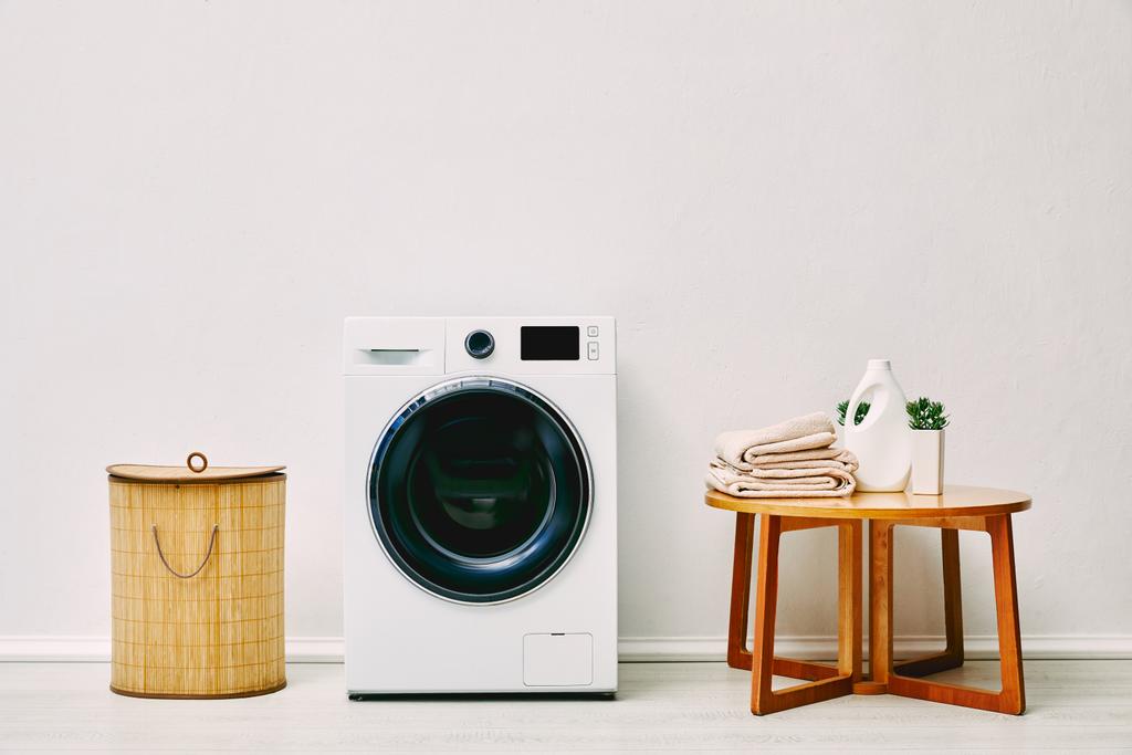 máquina de lavar roupa moderna perto de cesta de lavanderia, mesa de café com toalhas, garrafa de detergente e planta no banheiro
  - Foto, Imagem